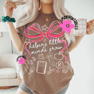 May include: A brown t-shirt with a large pink and red bow design and the phrase "helping little minds grow" in white script. The shirt also features white illustrations of school supplies, including an apple, notebook, and calculator.