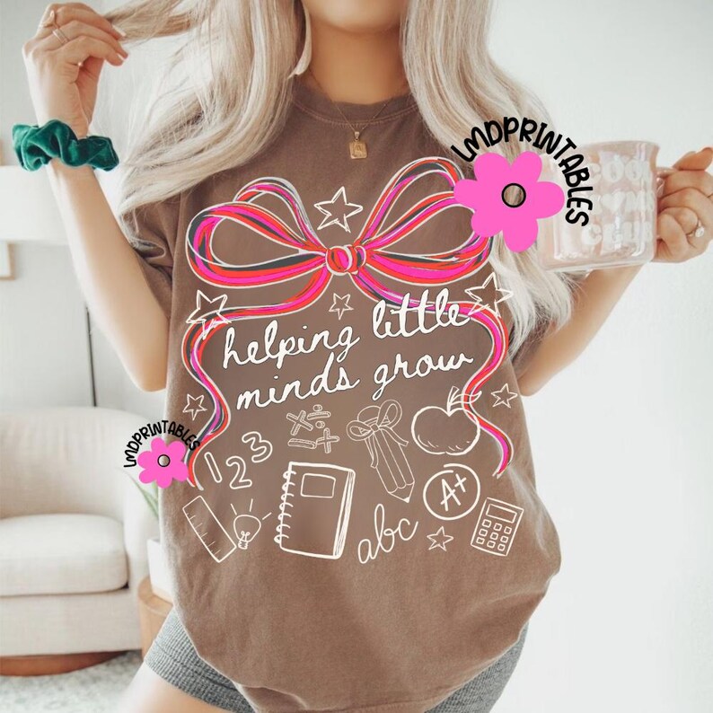 May include: A brown t-shirt with a large pink and red bow design and the phrase "helping little minds grow" in white script. The shirt also features white illustrations of school supplies, including an apple, notebook, and calculator.