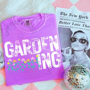 May include: A lilac-colored t-shirt with the word "GARDENING" in large white letters. Below the text is a row of colorful flowers. A newspaper with the headline "The New York" is in the background.