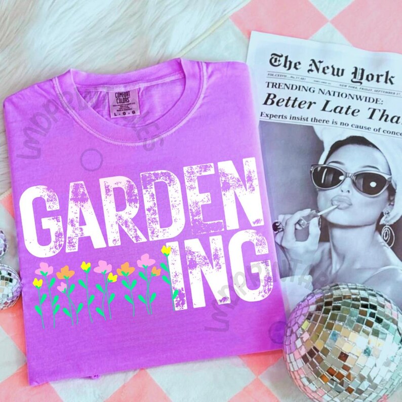 May include: A lilac-colored t-shirt with the word "GARDENING" in large white letters. Below the text is a row of colorful flowers. A newspaper with the headline "The New York" is in the background.