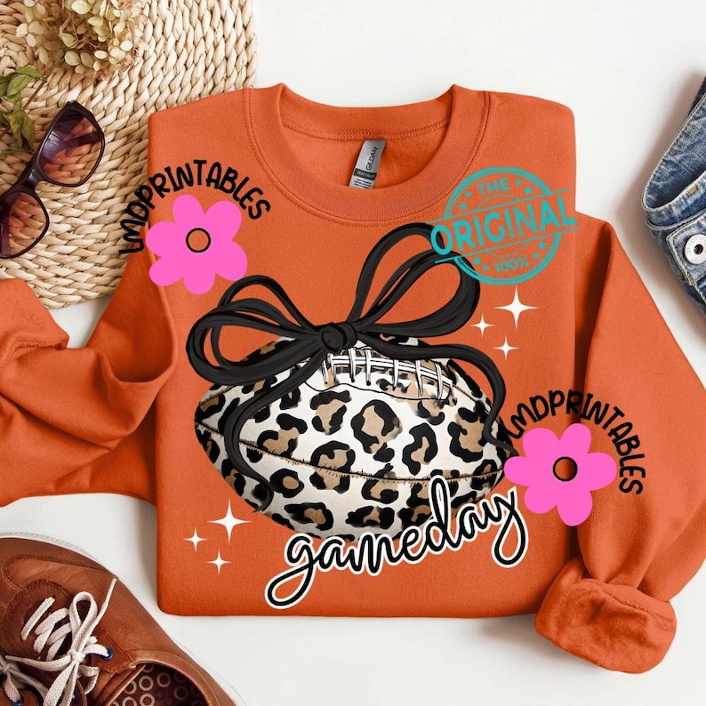 May include: An orange sweatshirt with a leopard print football design and the word "gameday" in a script font. The football is tied with a black bow. The text "MDPrintables" and a pink flower are on the left and right sides.