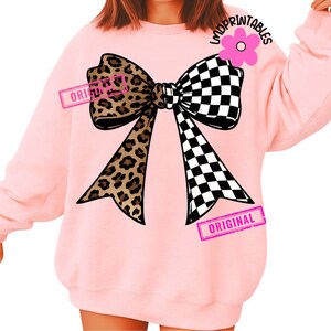 May include: Pink sweatshirt featuring a large bow design. The bow is split, with a leopard print on one side and a black and white checkered pattern on the other. The word "ORIGINAL" is stamped on the bow in pink.