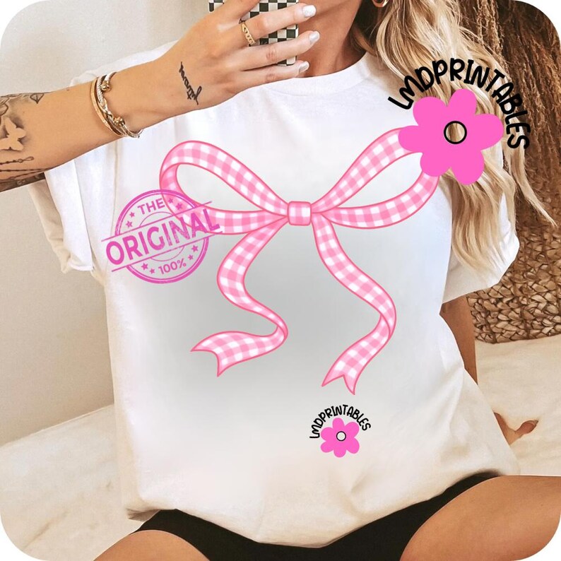 May include: White t-shirt featuring a large pink gingham bow design. The bow is centered on the shirt and has a ribbon-like appearance. The text "THE ORIGINAL 100%" is printed on a pink circular stamp. The shirt also has the text "LMDPRINTABLES" and a pink flower.