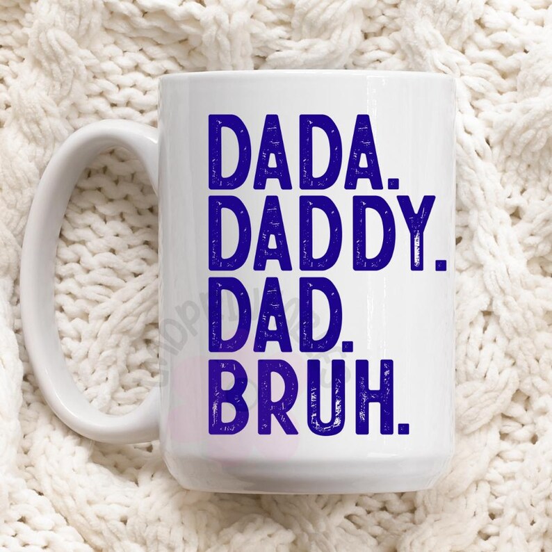 May include: White ceramic mug with the words "DADA. DADDY. DAD. BRUH." in dark blue, stacked vertically. The mug has a large handle and is set against a white, textured background.