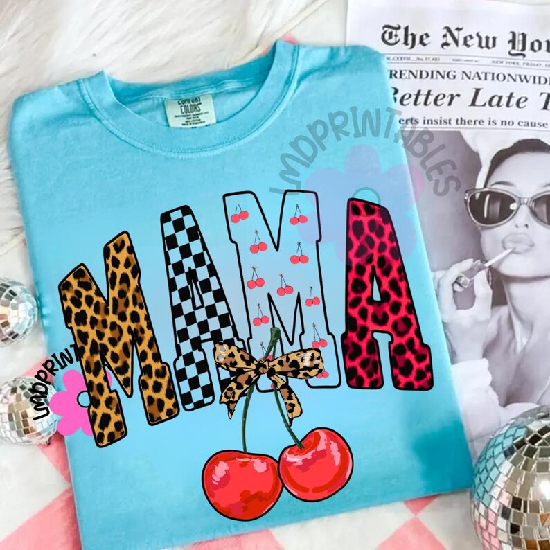 May include: A light blue t-shirt with the word "MAMA" in a mix of leopard print, checkered, cherry, and leopard print patterns. Two red cherries with a leopard print bow are at the bottom. The shirt has a crew neck.