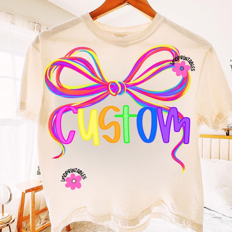 May include: Cream-colored t-shirt with a large, colorful bow design and the word "CUSTOM" in rainbow hues. The shirt features a round neckline and short sleeves. The design includes the text "UNDPRINTABLES" and a pink flower.