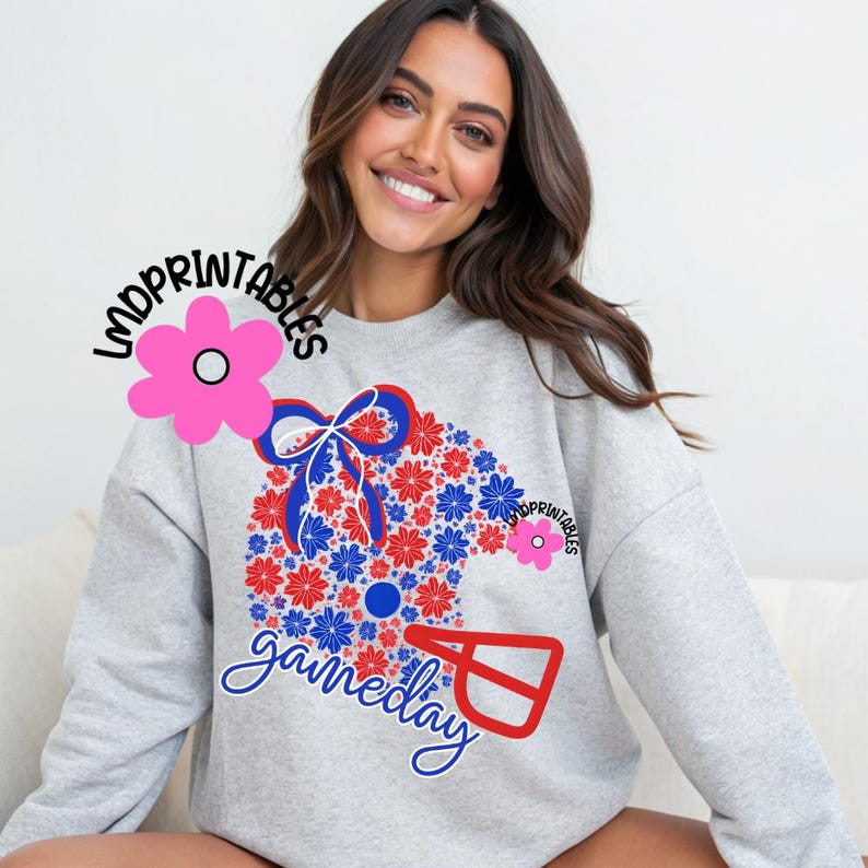 May include: A light gray sweatshirt featuring a floral design in red, white, and blue, with the word "gameday" in script. The design includes a football helmet shape filled with flowers and a bow.