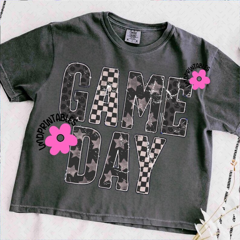 May include: Dark grey t-shirt with the words "GAME DAY" in a distressed, outlined font. The letters feature patterns like stars, leopard print, and checkered designs. A pink flower graphic is on the shirt.