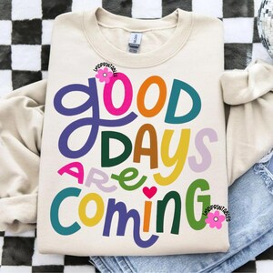 Może przedstawiać: Kremowa bluza z napisem "GOOD DAYS ARE COMING" w kolorowych, zabawnych literach. Projekt zawiera r&oacute;żowe serce i kwiatowe akcenty. Swobodny, wygodny top do noszenia na co dzień.