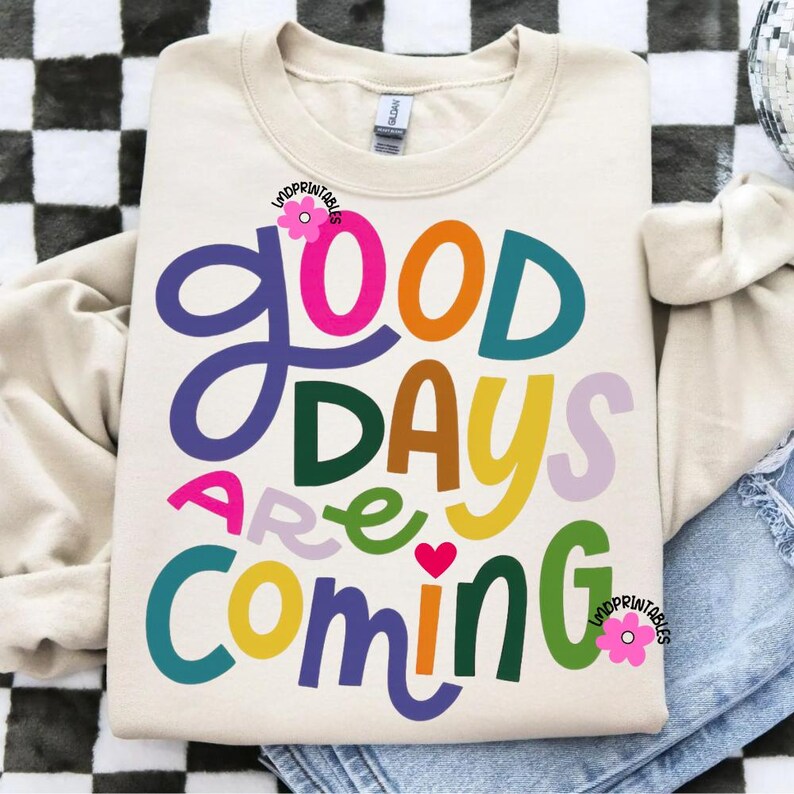 Może przedstawiać: Kremowa bluza z napisem "GOOD DAYS ARE COMING" w kolorowych, zabawnych literach. Projekt zawiera r&oacute;żowe serce i kwiatowe akcenty. Swobodny, wygodny top do noszenia na co dzień.