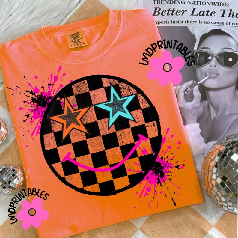 May include: An orange Comfort Colors sweatshirt with a smiley face design. The design includes a black and white chequered circle with orange and teal stars, a pink smile, and pink paint splatters. The text "LMDPRINTABLES" is also visible.