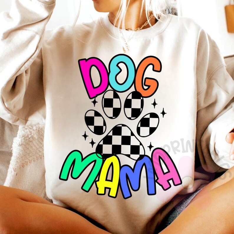 May include: Cream-colored sweatshirt with a colorful graphic. The graphic features the words "DOG MAMA" in bright, bubble letters, with a paw print design in the center. The paw print has a black and white checkered pattern.