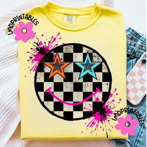 May include: A yellow t-shirt featuring a smiley face design. The smiley face has a black and white chequered background with orange and blue star eyes and a pink smile. Pink paint splatters and the text "LNDPRINTABLES" are also on the shirt.