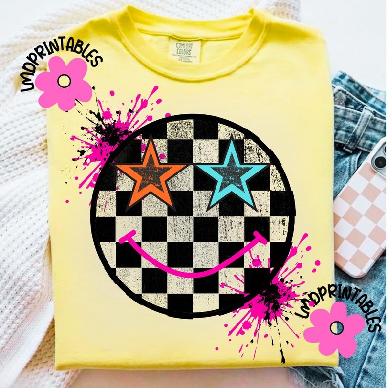 May include: A yellow t-shirt featuring a smiley face design. The smiley face has a black and white chequered background with orange and blue star eyes and a pink smile. Pink paint splatters and the text "LNDPRINTABLES" are also on the shirt.