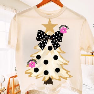 May include: Cream-colored t-shirt featuring a Christmas tree design. The tree is adorned with a black and white polka dot bow, black circles, and a gold star. The shirt is hanging on a wooden hanger.
