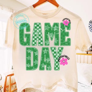 May include: Cream-colored t-shirt with the words "GAME DAY" in green, featuring a checkered pattern and star-filled letters. The shirt has a distressed look, with a pink flower graphic and the word "UNDPRINTABLES".