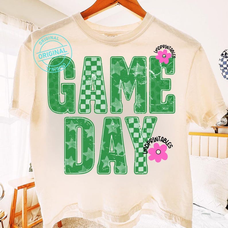 May include: Cream-colored t-shirt with the words "GAME DAY" in green, featuring a checkered pattern and star-filled letters. The shirt has a distressed look, with a pink flower graphic and the word "UNDPRINTABLES".