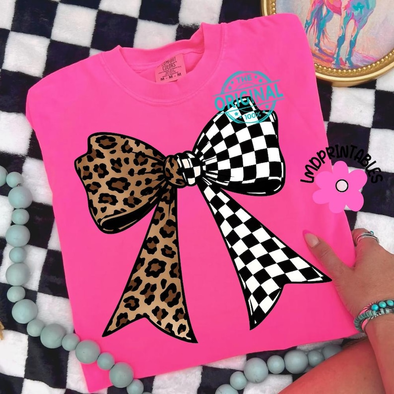 May include: A bright pink t-shirt featuring a large bow design. The bow is split, with a leopard print on the left and a black and white checkered pattern on the right. The text "THE ORIGINAL 100%" is printed on the top right.