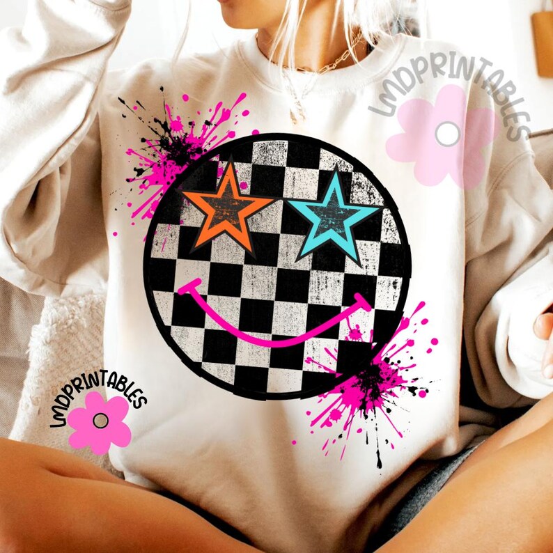 May include: A cream-coloured sweatshirt with a large graphic of a smiley face. The face has a chequered pattern, orange and blue stars for eyes, and a pink smile. Pink and black paint splatters surround the graphic. The text "LMDPRINTABLES" is printed on the shirt.