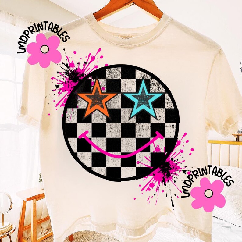 May include: A cream-coloured t-shirt with a smiley face design. The smiley face has a black and white chequered background, orange and blue star eyes, and a pink smile. Pink paint splatters and the text "IMPRINTABLES" are also visible.