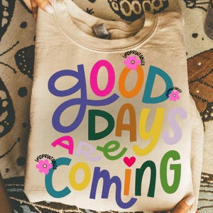 Może przedstawiać: Beżowa bluza z napisem "Good Days Are Coming" w kolorowych, zabawnych literach. Projekt zawiera r&oacute;żowy, pomarańczowy, niebieski, zielony, ż&oacute;łty i fioletowy tekst, z małym r&oacute;żowym sercem i kwiatowymi akcentami.