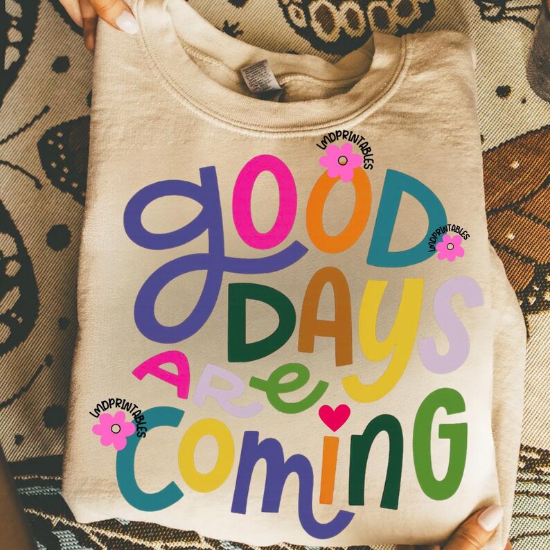 Może przedstawiać: Beżowa bluza z napisem "Good Days Are Coming" w kolorowych, zabawnych literach. Projekt zawiera r&oacute;żowy, pomarańczowy, niebieski, zielony, ż&oacute;łty i fioletowy tekst, z małym r&oacute;żowym sercem i kwiatowymi akcentami.