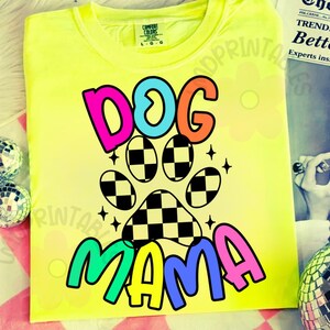 May include: A neon yellow t-shirt with a colorful graphic design. The design features the words "DOG MAMA" in vibrant colors, with a paw print in the center. The paw print has a checkered pattern. The shirt is made of a soft material.