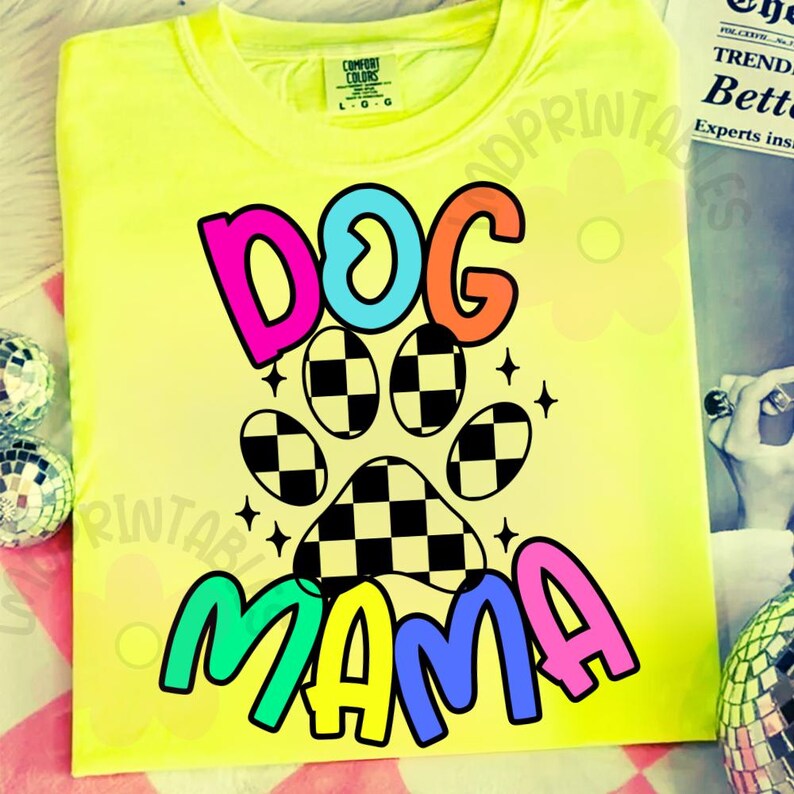 May include: A neon yellow t-shirt with a colorful graphic design. The design features the words "DOG MAMA" in vibrant colors, with a paw print in the center. The paw print has a checkered pattern. The shirt is made of a soft material.