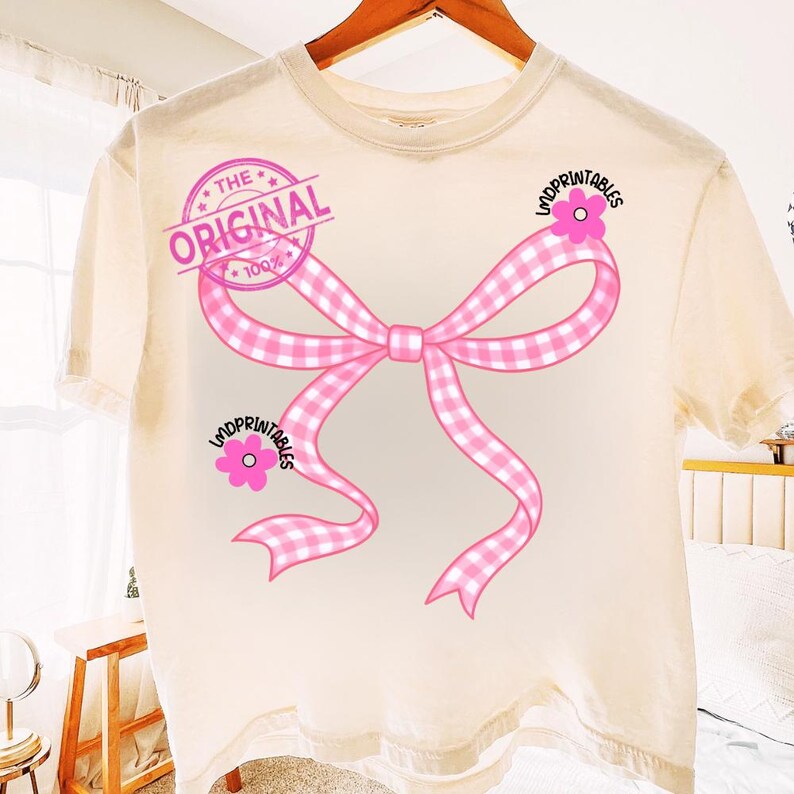 May include: Cream-colored t-shirt featuring a large pink bow with a gingham pattern. The bow design includes the text "THE ORIGINAL" in a pink stamp and the text "LMDPRINTABLES" with a pink flower on the shirt.