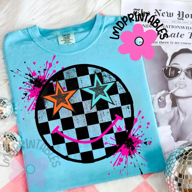 May include: A pale blue t-shirt with a black and white chequered circle design, featuring orange and turquoise stars and a pink smiley face. The shirt has pink and black paint splatters, and the text "IMPRINTABLES" and a flower graphic.