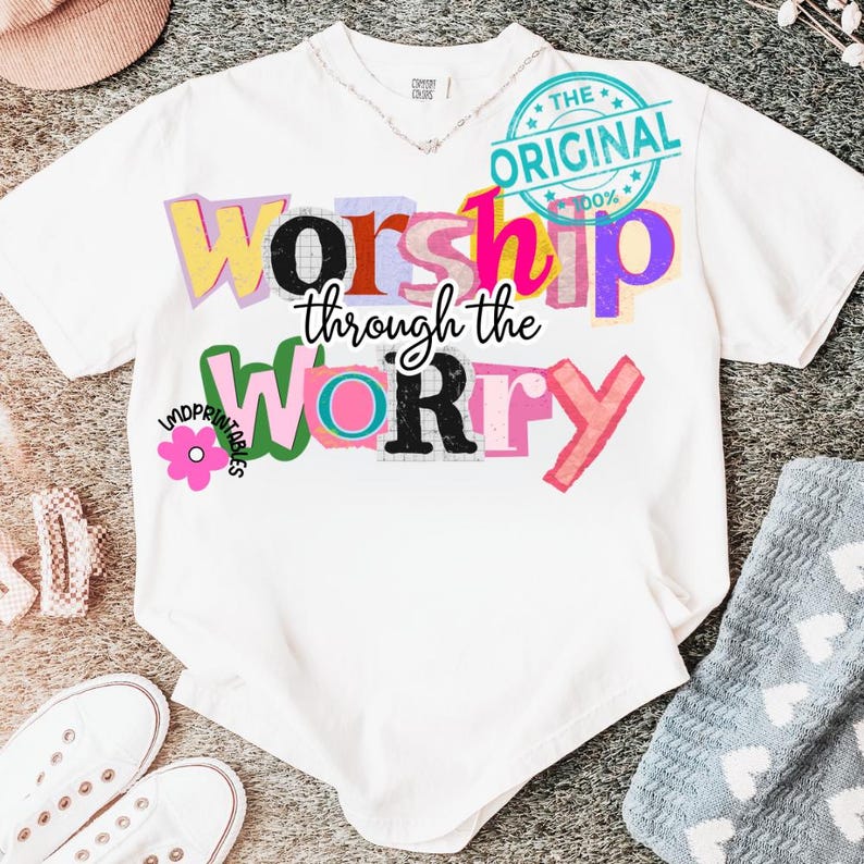 May include: White t-shirt with the words "Worship through the Worry" in colorful, layered text. The design includes a flower graphic and a "The Original" stamp. The shirt has a V-neck and short sleeves.