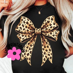 May include: Black t-shirt featuring a large bow design with a leopard print pattern. The bow is centered on the shirt and has a detailed, realistic appearance. The shirt also has a pink flower graphic and the text "LOVE PRINTABLES".