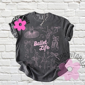 May include: Dark gray t-shirt with a pink ballet-themed design. The shirt features the text "Ballet Life" and illustrations of ballet dancers, tutus, bows, and pointe shoes. A silver necklace is visible.