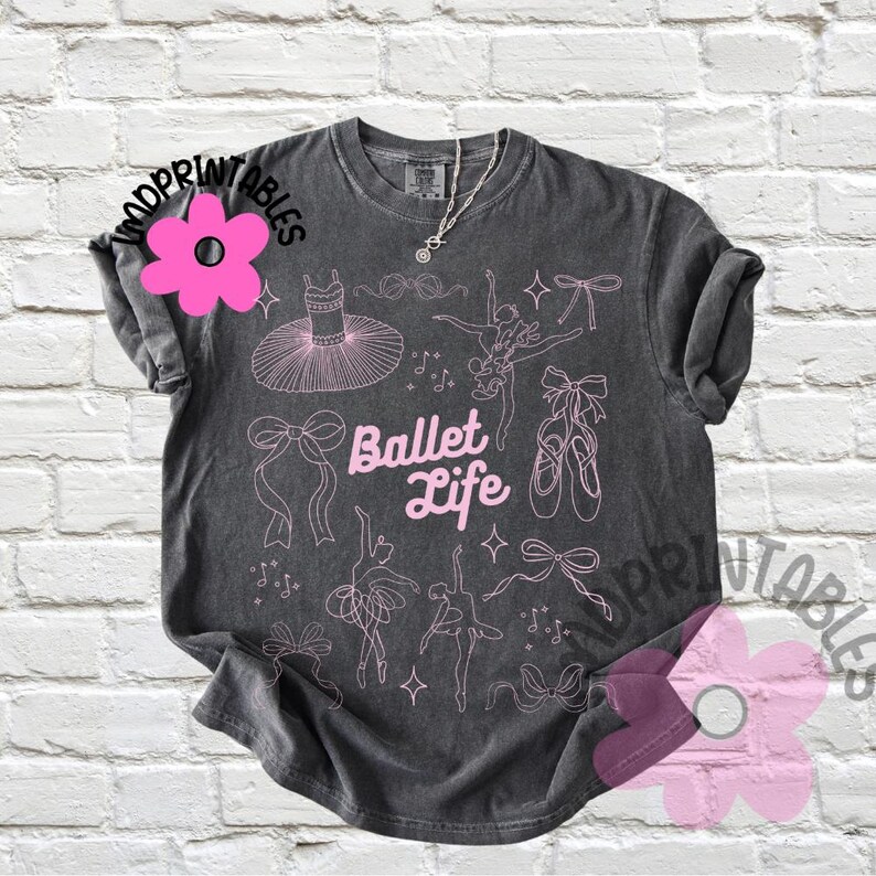 May include: Dark gray t-shirt with a pink ballet-themed design. The shirt features the text "Ballet Life" and illustrations of ballet dancers, tutus, bows, and pointe shoes. A silver necklace is visible.