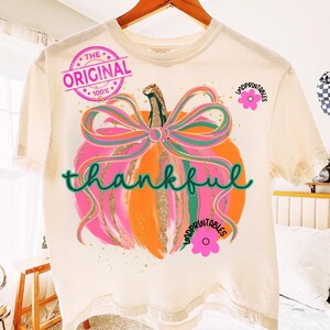 Peut inclure: T-shirt cr&egrave;me avec un motif de citrouille color&eacute;e. La citrouille est rose et orange avec un n&oelig;ud vert et rose. Le mot "thankful" est &eacute;crit en vert sur la citrouille. Le t-shirt porte &eacute;galement le texte "THE ORIGINAL 100%" et un motif de fleur rose.