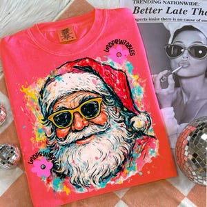 May include: A coral sweatshirt with a colourful graphic of Santa Claus wearing yellow sunglasses. The Santa illustration is surrounded by paint splatters in various colours. The text "LMDPRINTABLES" is printed on the shirt.
