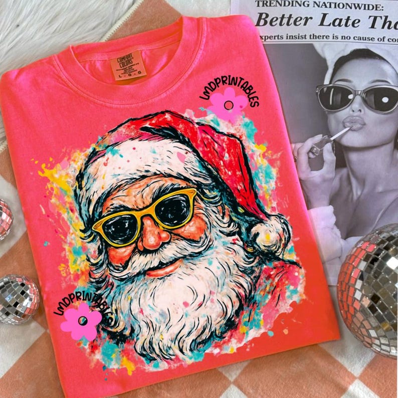May include: A coral sweatshirt with a colourful graphic of Santa Claus wearing yellow sunglasses. The Santa illustration is surrounded by paint splatters in various colours. The text "LMDPRINTABLES" is printed on the shirt.
