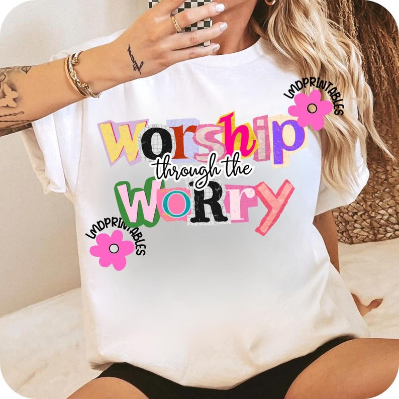 May include: White t-shirt with the text "Worship through the Worry" in colorful, block letters. The word "Worship" is in yellow, pink, and purple. The word "Worry" is in green, black, and pink. Pink flower graphics are on the shirt.