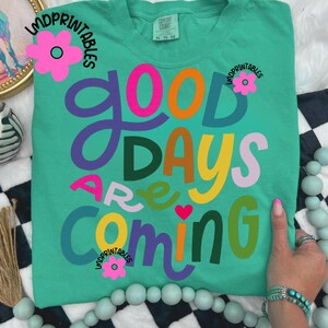 Może przedstawiać: Turkusowa koszulka Comfort Colors z napisem "Good Days Are Coming" w kolorowych, fantazyjnych literach. Widoczne są r&oacute;wnież r&oacute;żowe kwiaty i tekst "LMDPrintables". Koszulka ma luźny kr&oacute;j.