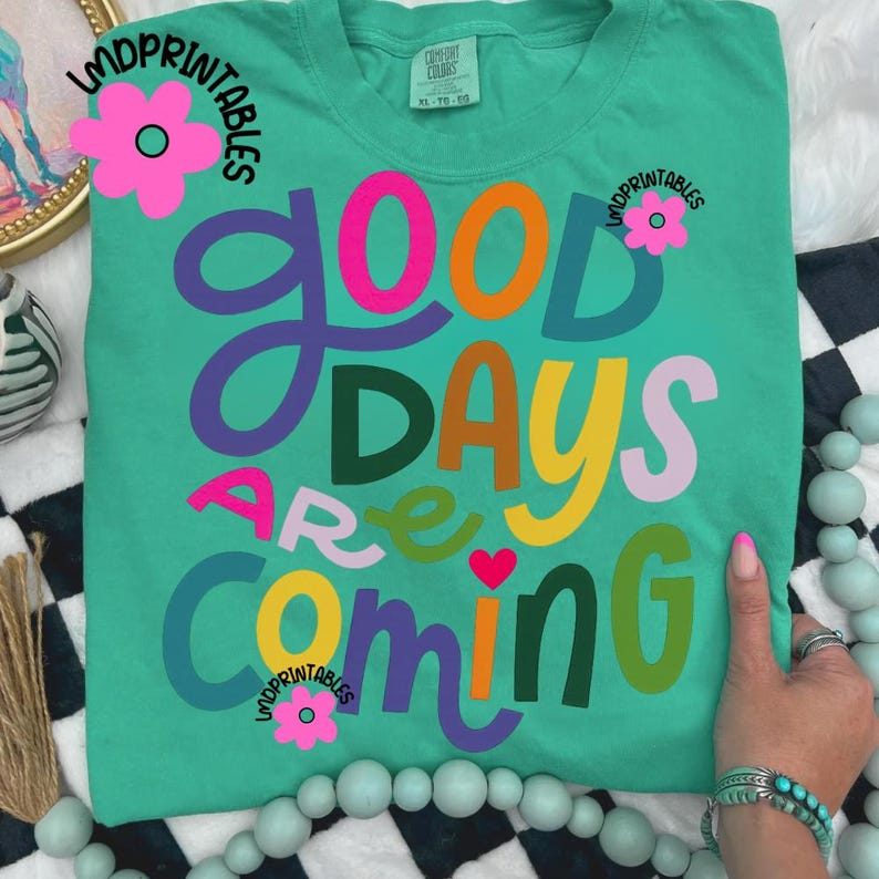 Może przedstawiać: Turkusowa koszulka Comfort Colors z napisem "Good Days Are Coming" w kolorowych, fantazyjnych literach. Widoczne są r&oacute;wnież r&oacute;żowe kwiaty i tekst "LMDPrintables". Koszulka ma luźny kr&oacute;j.