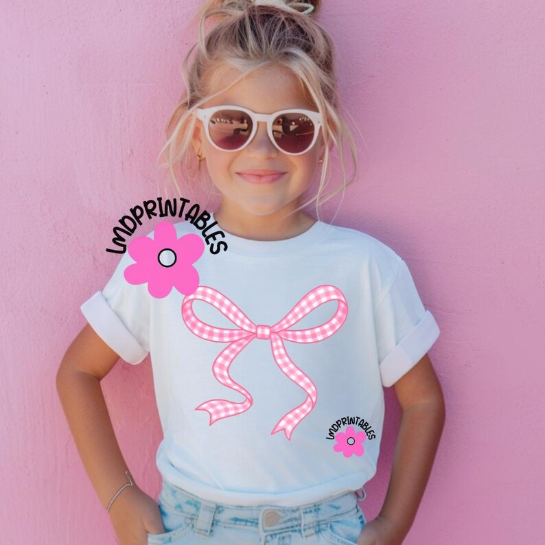 May include: White t-shirt featuring a pink gingham bow and two pink flowers. The shirt has rolled-up sleeves and the text "LMDPRINTABLES" is printed on the shirt. The background is a solid pink color.