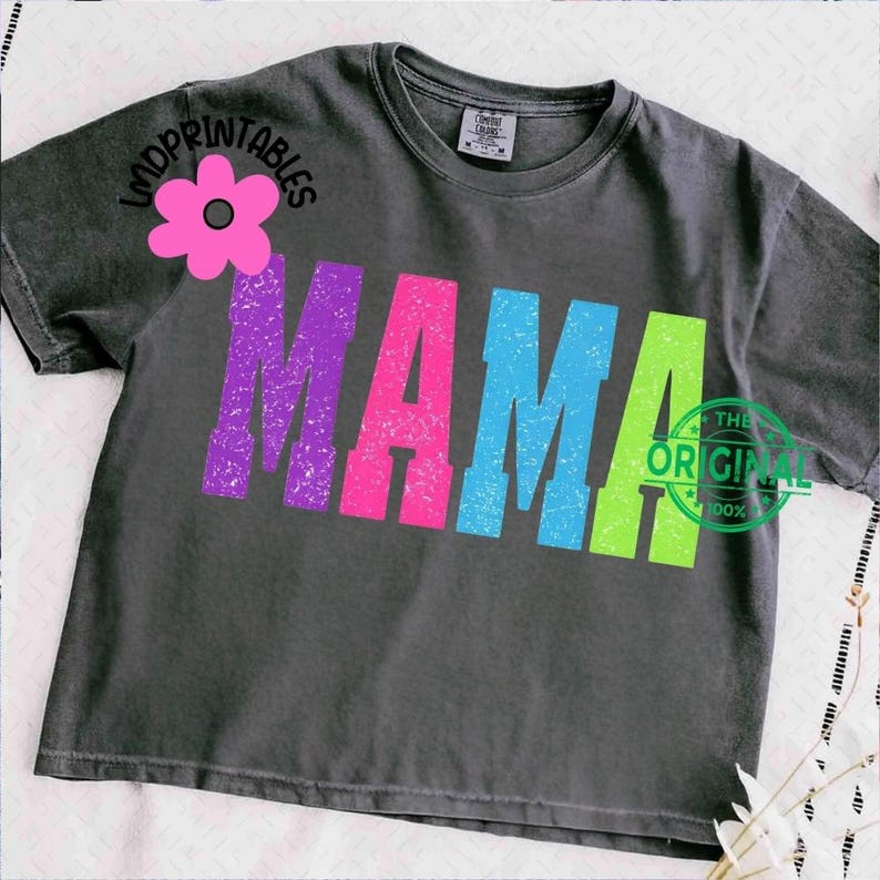 Pu&ograve; includere: T-shirt grigio scuro con la scritta "MAMA" a grandi lettere colorate e consumate. Le lettere sono viola, rosa, blu e verdi. Una grafica floreale rosa e la scritta "LMDPRINTABLES" sono sulla spalla sinistra. Il testo "THE ORIGINAL 100%" &egrave; a destra.