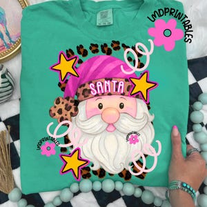Op de afbeelding: Petrolkleurig T-shirt met een afbeelding van de Kerstman. De Kerstman draagt een roze hoed met het woord "SANTA" en een luipaardprint. Het ontwerp bevat gele sterren, roze bloemen en krullen. De tekst "LMDPRINTABLES" is ook zichtbaar.