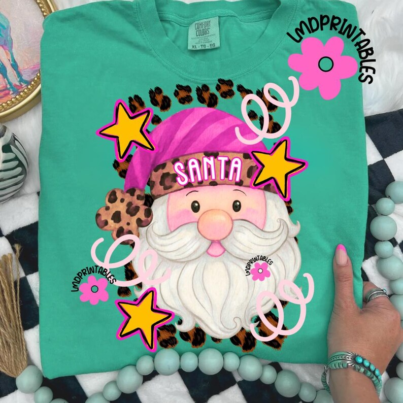 Op de afbeelding: Petrolkleurig T-shirt met een afbeelding van de Kerstman. De Kerstman draagt een roze hoed met het woord "SANTA" en een luipaardprint. Het ontwerp bevat gele sterren, roze bloemen en krullen. De tekst "LMDPRINTABLES" is ook zichtbaar.