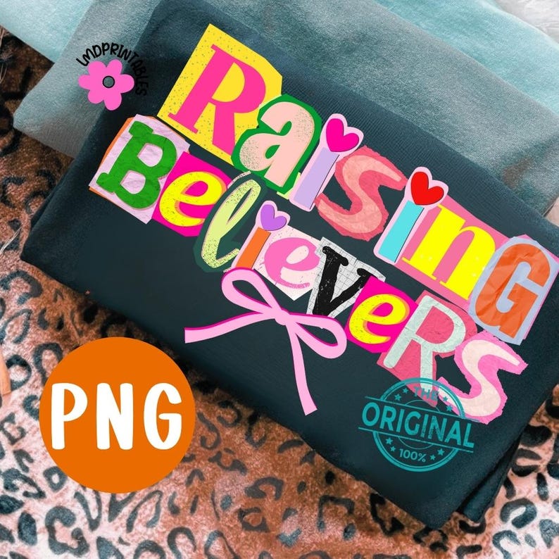 May include: A dark teal t-shirt with the words "Raising Believers" in colorful, cut-out style letters. A pink bow is below the text. The image also includes a PNG logo and a "Original 100%" stamp.