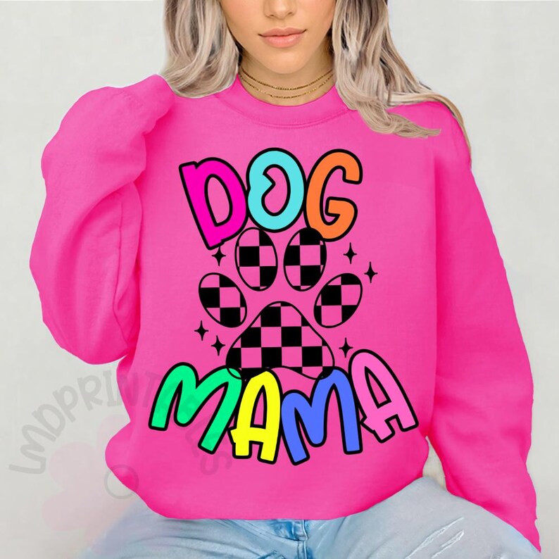 May include: A bright pink sweatshirt with the words "DOG MAMA" in colorful, bubble letters. A paw print with a checkered pattern is in the center. The sweatshirt is a crew neck style.