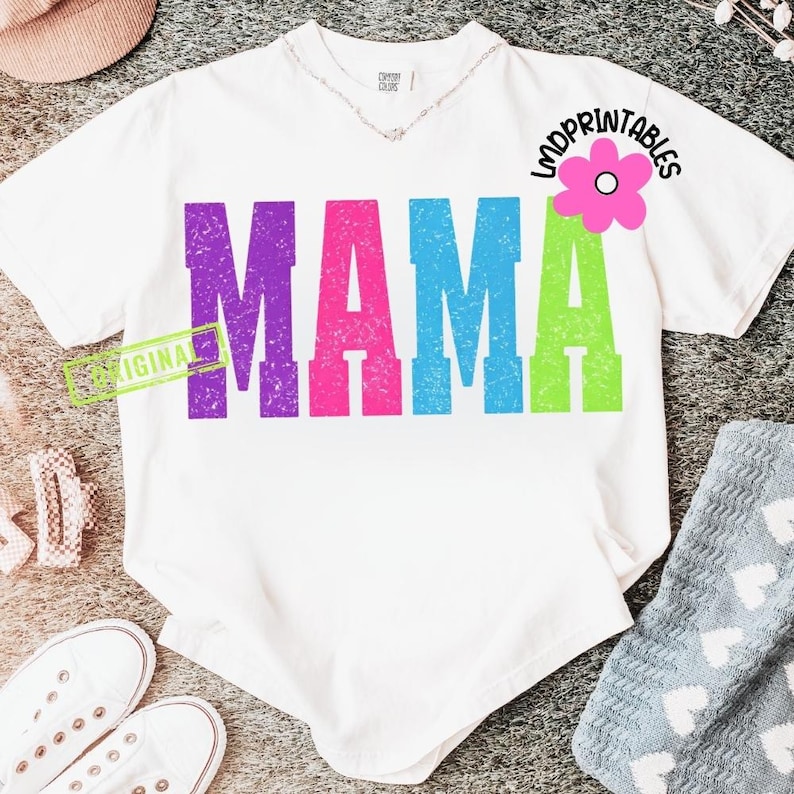 Pu&ograve; includere: T-shirt bianca con la scritta "MAMA" in lettere a blocchi viola, rosa, blu e verdi. Una grafica floreale rosa con la scritta "LMDPrintables" si trova in alto a destra. La parola "ORIGINAL" &egrave; stampata su un rettangolo verde.