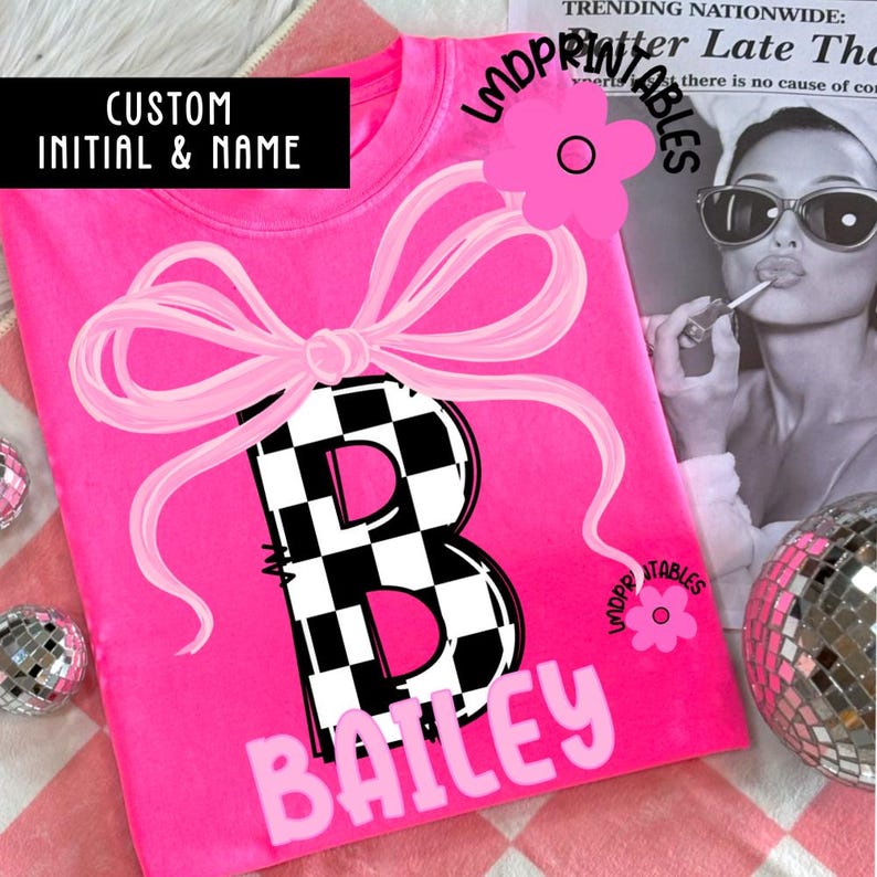 May include: A hot pink t-shirt featuring a large letter "B" with a black and white checkered pattern, a pink bow, and the name "Bailey" in white. The text "CUSTOM INITIAL & NAME" is in a black rectangle.