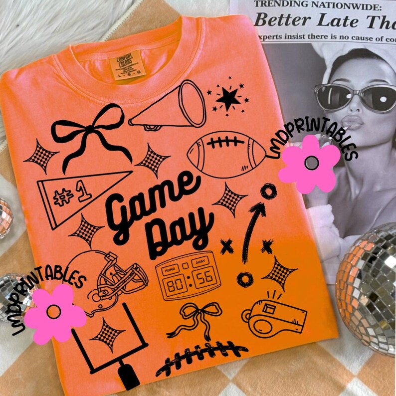 May include: Orange t-shirt with black graphic illustrations of football-related items, including a football, helmet, and the words "Game Day." The shirt also features a scoreboard, a whistle, and a bow.