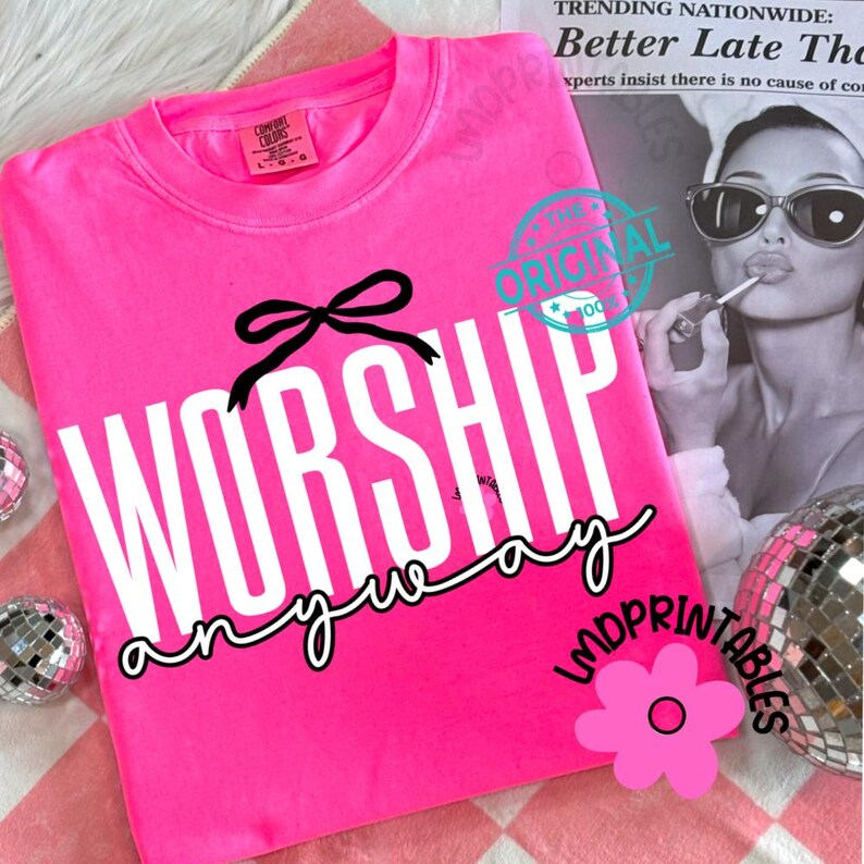 May include: A vibrant pink sweatshirt featuring the text "WORSHIP anyway" in white, with a black bow graphic. The crew neck top is a Comfort Colors brand. The design is centrally placed on the front of the garment.